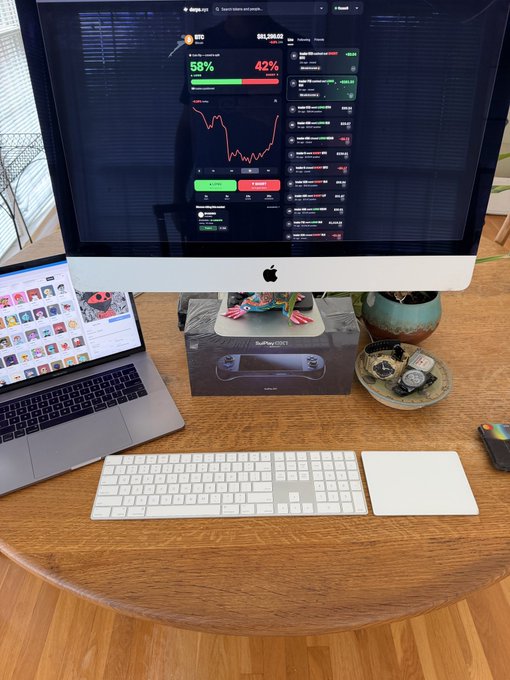 A wooden desk setup features an iMac displaying crypto trading charts with BTC at 58% and 42%, alongside a MacBook laptop, a white keyboard and trackpad, and a Steam Deck box.