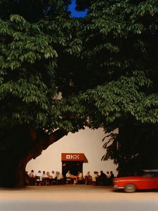 A nighttime outdoor café scene beneath a massive, leafy tree, where patrons sit at tables under a sign reading "OKX," while a red vintage car speeds past. The image evokes the post's message of a world just waking up while the 24/7 crypto market keeps moving — OKX operating continuously even as day transitions to night.