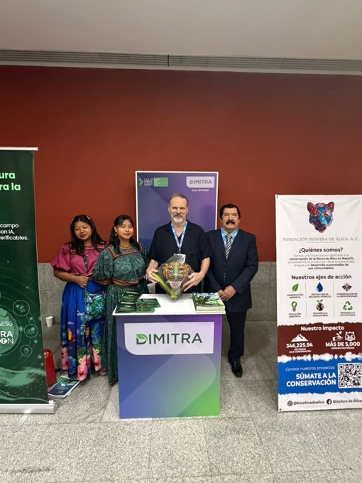 The image shows a group of people standing around a booth at an event, with a sign that says "DMTR" on it. The booth appears to be promoting a product or service related to carbon credits. There are four individuals present, including two men and two women. One man is holding up a trophy, while the other three are posing for a photo in front of the booth. A banner can also be seen behind them, which has information about the event and the company they represent.