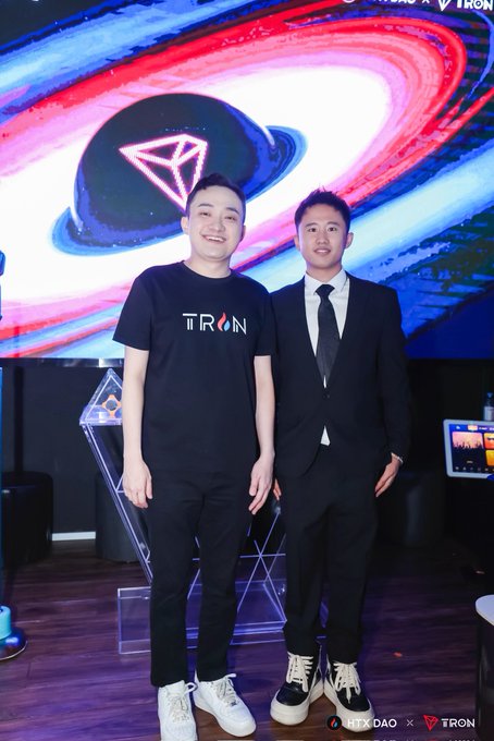 Two men pose together in front of a TRON-branded backdrop featuring the iconic diamond logo against a colorful cosmic design; one wears a black "TRON" t-shirt and white sneakers, while the other is dressed in a dark suit with a tie, with the "HTX DAO × TRON" logo visible at the bottom right. The image relates to the post as a celebratory birthday moment honoring Justin Sun (brother Sun), the founder of TRON, with the attendee likely referencing their commitment to continue working alongside him in the crypto industry.