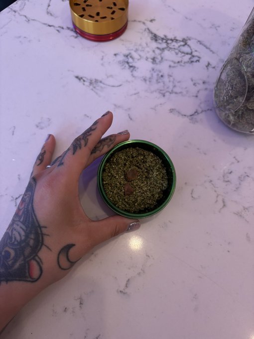 A tattooed hand holds a green tin containing a green herbal substance (likely kush) on a white marble-patterned countertop, with a glass jar of similar material visible in the background.