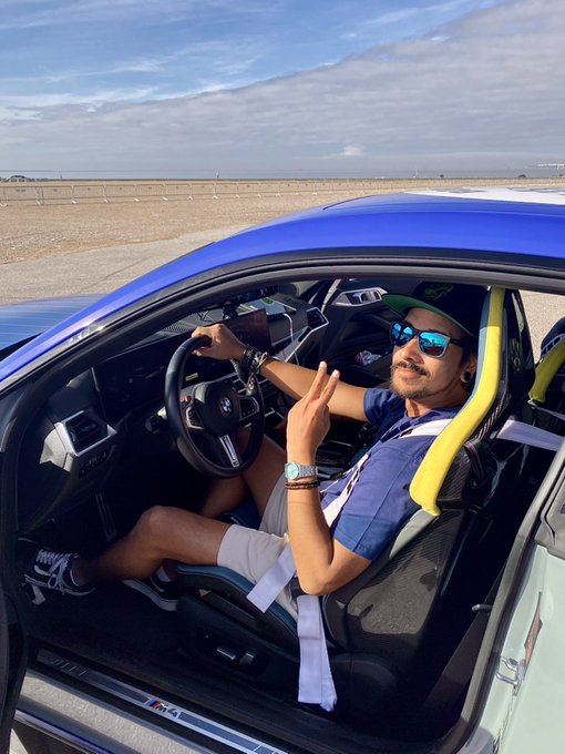 A young man wearing sunglasses and a cap poses with a peace sign while seated inside a blue BMW M4, with the "M4" badge visible on the door sill. The post "Gm Legends" suggests this is a celebratory or motivational post, matching the confident, victorious gesture against the open landscape backdrop.