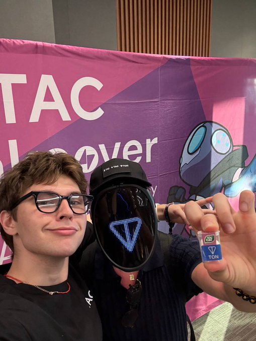 A young man wearing glasses taking a selfie with someone wearing a robotic helmet displaying a glowing blue 'T' logo at a TON event, holding a TON branded item.