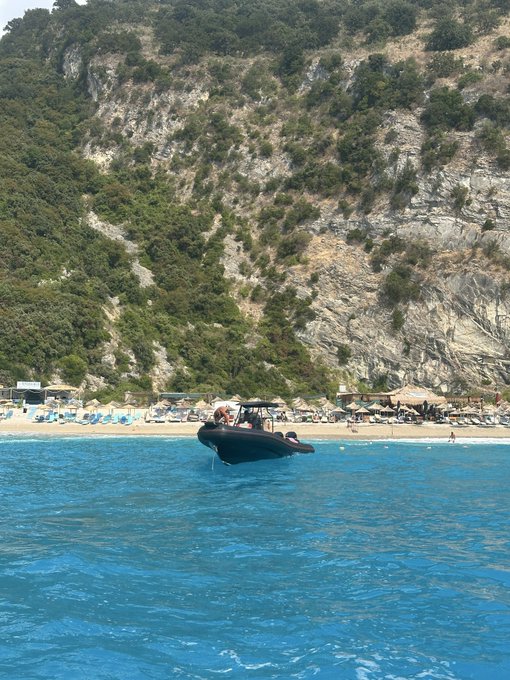 A small boat floats in bright turquoise water near a beach with umbrellas, backed by a towering rocky cliff covered in greenery.