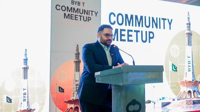 Farrukh Khan, Bybit's Pakistan Country Manager, delivers a speech at a podium during the Bybit Community Meetup in Lahore, with the event's branded backdrop and Pakistan flag imagery visible behind him. The caption aligns with the post's mention of Country Manager @farrukhkeyani addressing attendees at this inaugural Bybit South Asia community gathering.