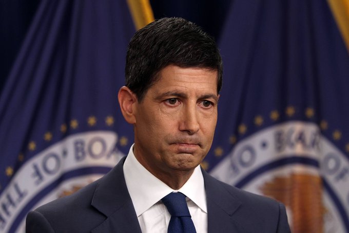 A serious-looking man in a dark suit and tie stands before a backdrop featuring seals of the Board of Governors of the Federal Reserve System.