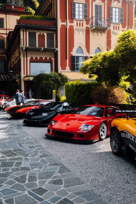 A scenic view of luxury sports cars including a red Ferrari and other vehicles parked on a stone-paved street in front of an ornate orange and beige Italian-style building with shutters and balconies.
