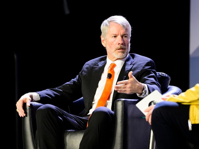 Michael Saylor, the chairman and co-founder of MicroStrategy (MSTR), is captured mid-speech at what appears to be a conference or panel discussion, gesturing with his hands while seated in a chair. The image visually accompanies his commentary on BIP-110 and Bitcoin consensus, as his serious, engaged expression and speaking posture reflect the assertive tone of his statement that "consensus is earned, not declared."