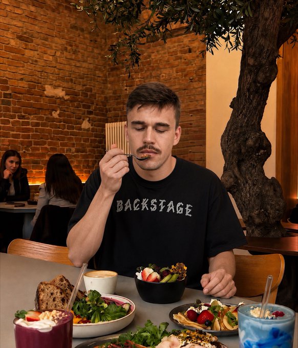 A young man with a mustache wearing a black 'BACKSTAGE' t-shirt sits at a restaurant table eating with a spoon, surrounded by an assortment of colorful healthy dishes including smoothies, salads, fruit bowls, and bread, with a brick wall and an indoor tree visible in the background.