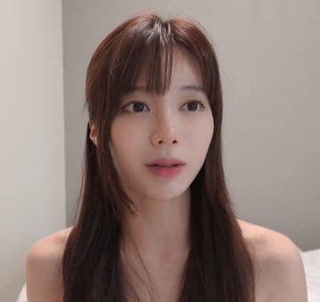 A close-up portrait of a young woman with long dark hair and bangs, looking directly at the camera with a soft expression against a plain beige background.