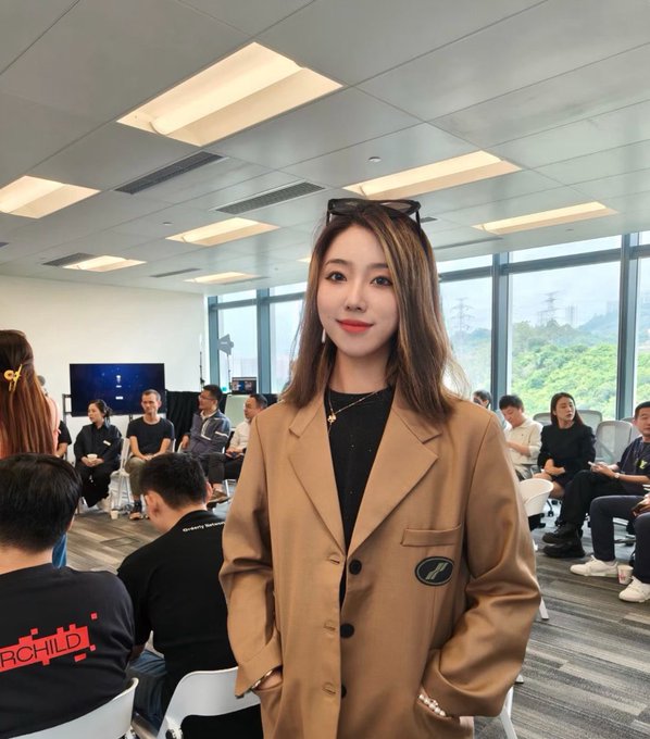 The image features a woman wearing a brown coat, standing in front of a group of people who are sitting down. She appears to be posing for a photo or participating in an event. The room has a white ceiling with multiple lights that illuminate the space. There is also a TV mounted on the wall near the center of the room.

The woman's attire includes a black top and sunglasses, which adds a stylish touch to her appearance. She seems to be enjoying herself as she poses for the picture or engages in conversation with others present in the room.