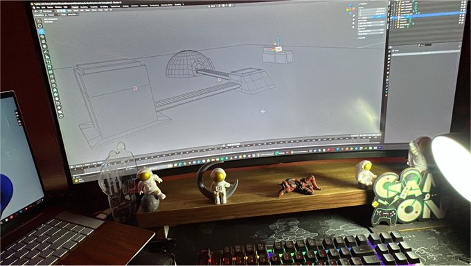 A workspace setup shows a large monitor displaying a 3D modeling software interface with geometric shapes, alongside a laptop, mechanical keyboard, and several small figurines on a wooden stand.