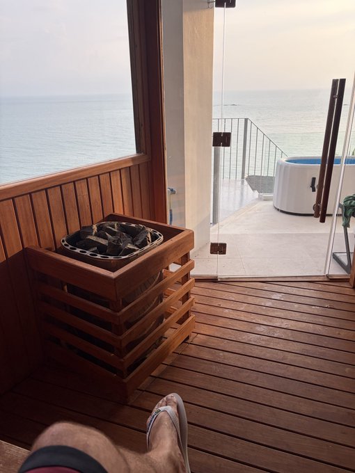 First-person view from a wooden deck overlooking the ocean, with feet in flip-flops visible, a wooden planter with rocks, and an outdoor hot tub through a glass door.