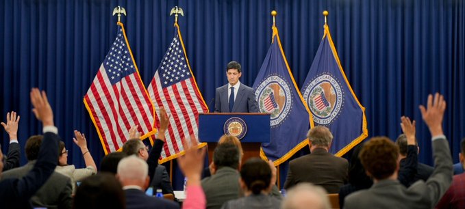 A man in a suit speaking at a podium bearing the presidential seal, with American flags behind him and an audience with raised hands in the foreground.