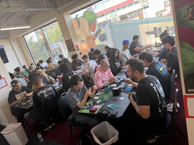 A crowded room full of people seated at long tables playing card games, likely a trading card game tournament, with large windows and street art visible in the background.