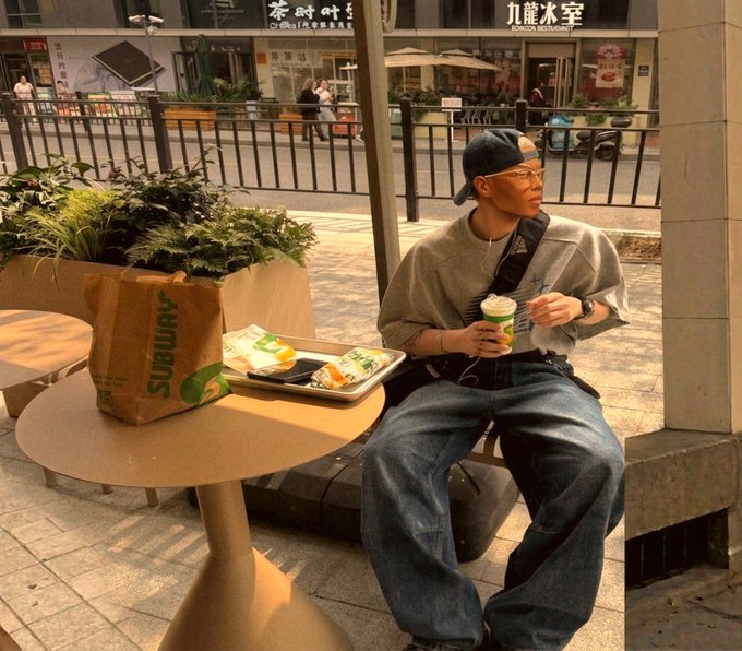 A young man in a gray sweatshirt, jeans, and a backward cap sits at a small table holding a Subway sandwich bag and drinks, with Chinese storefront signage visible across the street.