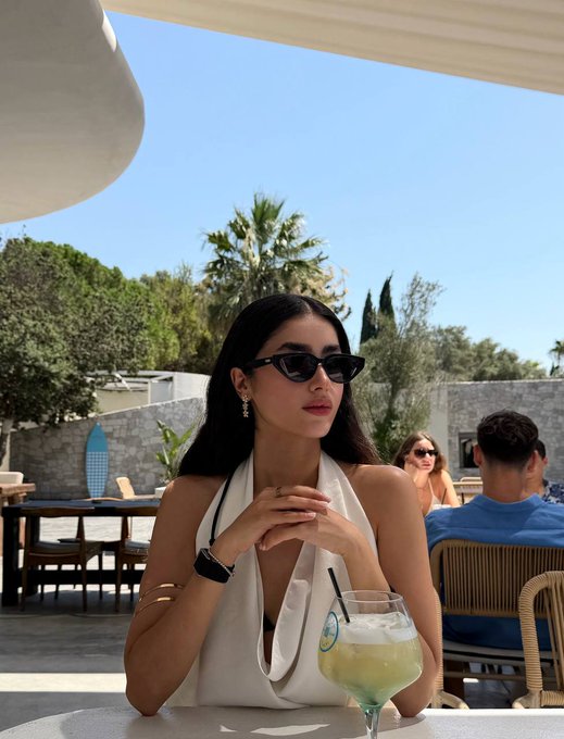 A young woman with long dark hair wearing cat-eye sunglasses and a white halter top, sitting at an outdoor restaurant table with a green cocktail, with palm trees and other diners visible in the background.