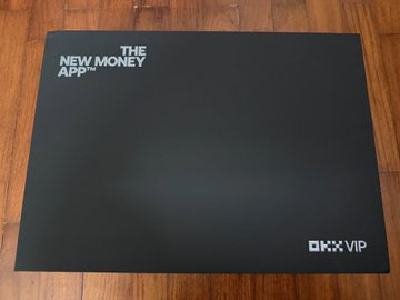 A black OKX VIP gift box labeled "THE NEW MONEY APP" sits on a wooden surface, corresponding to the post's mention of receiving an OKX VIP gift package.