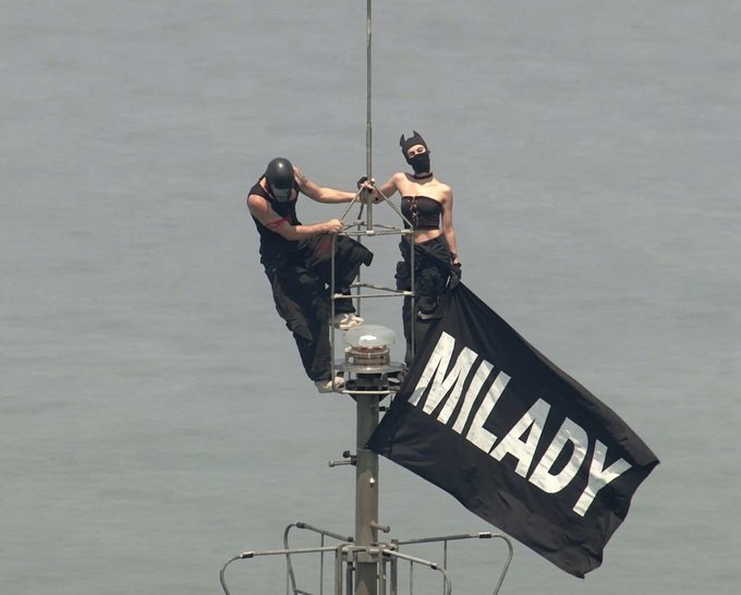 Two masked individuals in black attire perch atop a ship's mast against an open sea backdrop, unfurling a large black flag emblazoned with "MILADY" in bold white lettering—evoking a pirate-style declaration for the Milady meme coin.