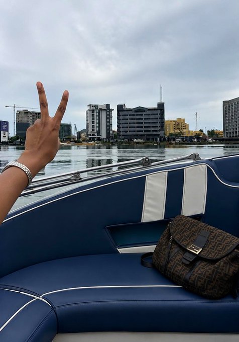 A hand making a peace sign on a boat with blue and white seats, with a brown Fendi bag resting beside it, overlooking a cityscape with buildings under a cloudy sky.