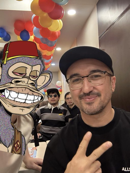 A man wearing glasses and a black cap poses with a peace sign next to a cardboard cutout of a cartoon monkey character with a fez.