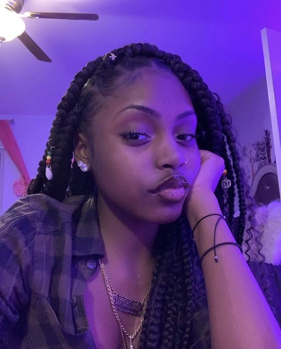 A close-up selfie of a young woman with long braids adorned with beads, wearing a nose ring, lip ring, and plaid shirt, bathed in purple lighting.