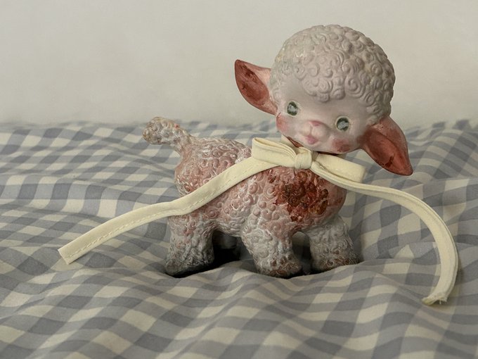 A vintage pink ceramic lamb figurine with curly textured wool and a cream-colored ribbon bow rests on a blue and white checkered cloth.