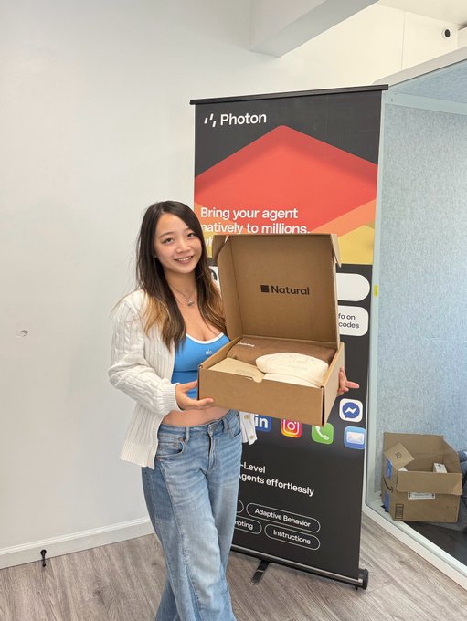 A smiling woman holds open a branded "Photon" and "Natural" box at a startup event, aligning with the post celebrating the company's raise and showcasing its agentic payment experience on iMessage via @PhotonHQ. The banner behind her reads "Bring your agent natively to millions," reinforcing the product's launch and partnership announcement.