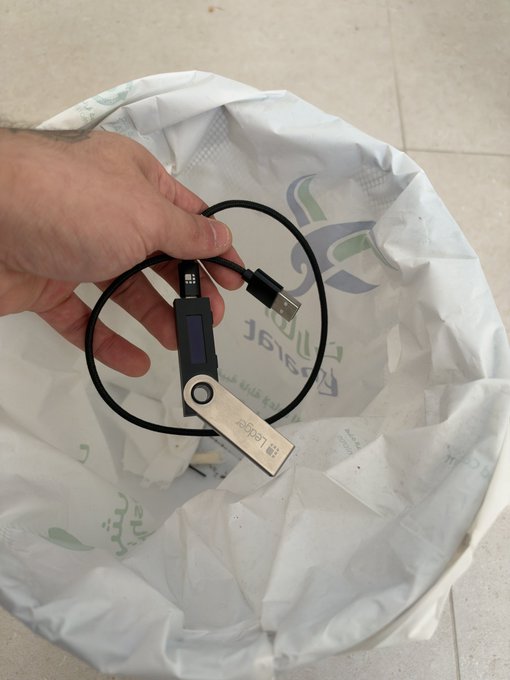 A hand holds a Ledger Nano S hardware wallet above a trash bin lined with a plastic bag, symbolically discarding traditional crypto storage in favor of a more secure hardware wallet solution.
