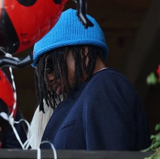 A person with long dreadlocks wearing a blue knit beanie and dark blue sweater looks down, partially obscured by balloons in the foreground.