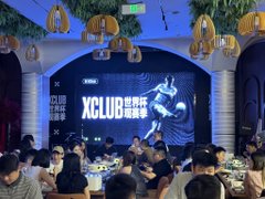 A crowded indoor event venue features a large screen displaying "XCLUB" and Chinese characters alongside a flying bull logo, with the "OKX" branding visible on a side banner. Attendees sit at tables in the foreground watching the brightly lit stage decorated with hanging wicker lanterns, reflecting the lively offline meetup described in the post.