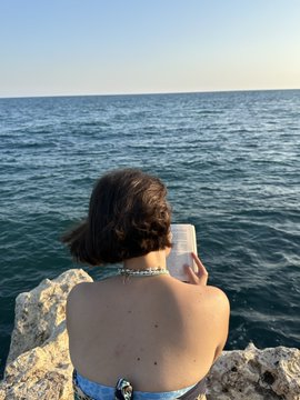 A woman with dark hair, seen from behind, sits on rocks by the sea while holding a drink.