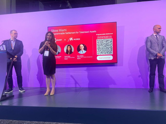 A woman speaks on stage at SWIFT's Sibos Miami symposium during a Quant x Murex presentation titled "Programmable Settlement for Tokenised Assets," flanked by two men. The screen displays headshots of speakers Launia Rule (Quant) and Maryam Bui (Murex) alongside a QR code for the "Programmable Settlement" demo, illustrating the live Delivery vs. Payment (DvP) demonstration mentioned in the accompanying post about $QNT.