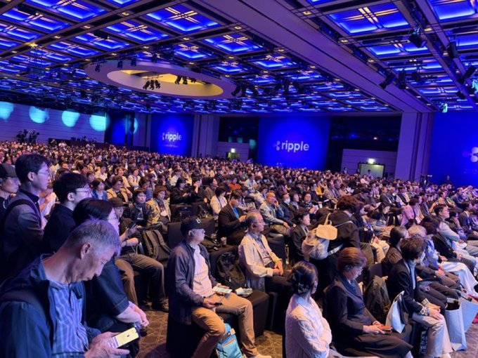 A large audience is seated in a modern conference hall with a blue-lit ceiling, watching a Ripple presentation displayed on screens.