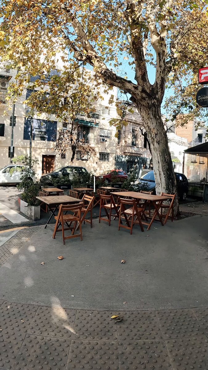 The image shows a street scene featuring an outdoor seating area with several wooden tables and chairs arranged under a large tree. The table settings include a pizza box that says "Bought pizza in Argentina with River satUSD Shop got ARS instantly Felt like any normal afternoon, pizza in hand and sorted at the counter". A car is parked nearby, adding to the urban atmosphere of the scene.

The image also includes some text elements such as a stop sign on the right side and a street lamp further down the road. The overall setting appears to be an outdoor cafe or restaurant with a relaxed ambiance, inviting passersby to enjoy their pizza in a comfortable environment.