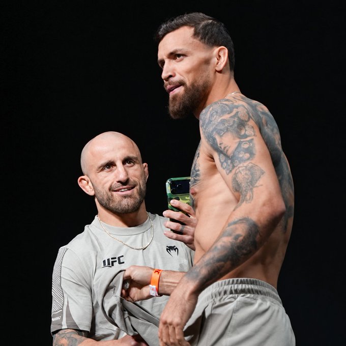 A man wearing a gray UFC Venum t-shirt smiles while holding up a smartphone displaying a green screen, standing beside a shirtless, heavily tattooed fighter. The image ties into a social media post celebrating a $14,181 bet placed on Mauricio Ruffy to defeat Arman Tsarukyan at UFC 331, with the fighter pictured likely being Tsarukyan during a weigh-in or pre-fight event.