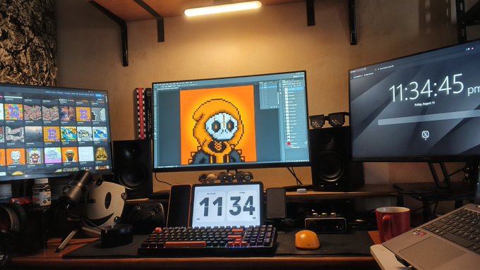 A dual-monitor desk setup displaying pixel art, with the left screen showing a gallery, the center monitor showing a yellow skull character, the right monitor showing the time '11:34:45 pm', and a digital clock showing '11 34' below.