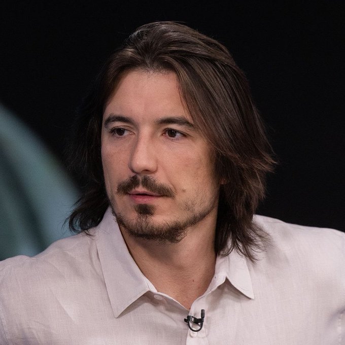A man with long brown hair and a goatee, wearing a white button-up shirt with a small microphone clipped to it, sits against a dark background during what appears to be a televised interview. The image appears unrelated to the post about Robinhood Chain's Ethereum revenue, as it simply shows a person being interviewed without any visible text, logos, or financial data.