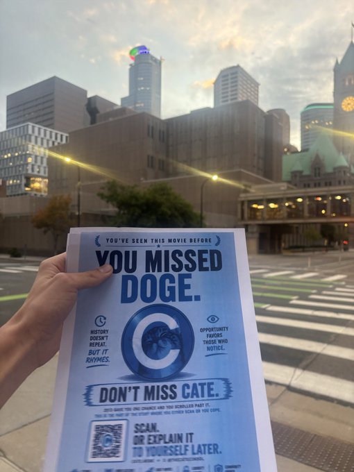 A hand holds up a low-toner blue $CATE flyer reading "YOU MISSED DOGE. DON'T MISS CATE" in downtown Minneapolis, matching the caption's note that the printer only had blue ink left while spreading the word about $CATE on the streets.