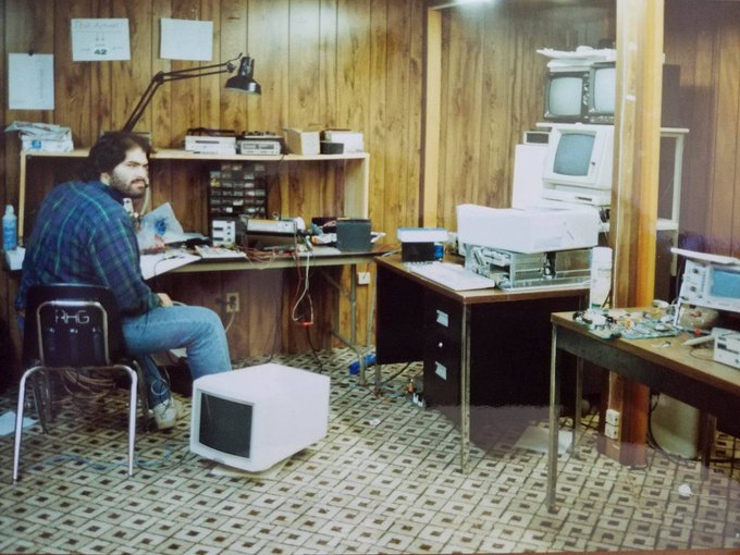A bearded man in a plaid shirt sits at a wood-paneled desk surrounded by vintage computer equipment, monitors, and electronic components in what appears to be a 1980s-era electronics repair shop. While the caption suggests a late-night work dedication theme related to the speaker's observations, the image itself simply depicts the man in his cluttered technical workspace, with no visible text identifying him or connecting to XRP.