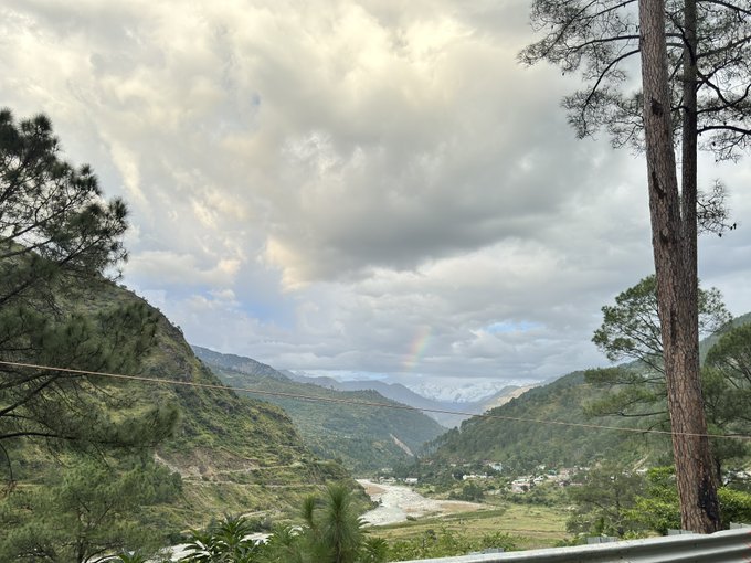 A scenic landscape view of a lush green mountain valley with a rainbow arching in the cloudy sky, framed by pine trees in the foreground.