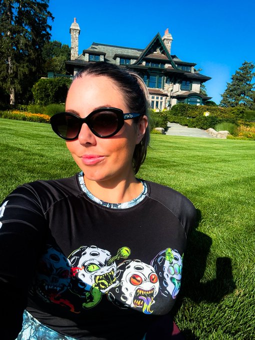 A woman wearing sunglasses and a black shirt with colorful alien/skeleton graphics takes a selfie in front of a large Victorian-style mansion on a sunny day.