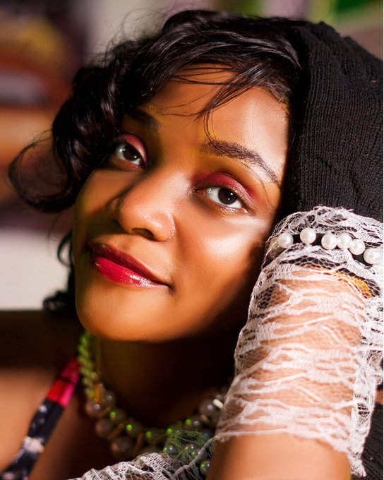 A radiant close-up portrait captures a woman with striking makeup—glossy red lips and warm-toned eyeshadow—posing beside her arm draped in a pearl-embellished lace sleeve and a black headwrap, embodying the stylish, creative "Ruby" celebrating her August birthday with bold self-expression.