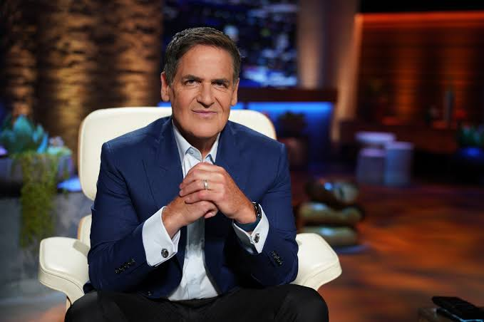 Businessman Mark Cuban sits in a white chair wearing a navy blue blazer over a white dress shirt, posing with his hands clasped in a studio setting.