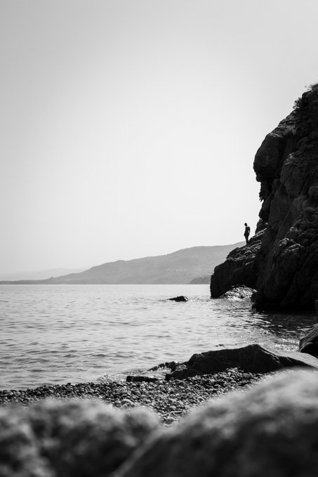 A black and white landscape photograph showing a small figure standing on rocky cliffs beside a calm body of water.