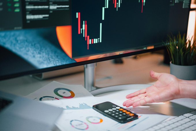 A workspace shows a hand reaching toward financial documents, a calculator, and a monitor displaying a red and green candlestick stock market chart.