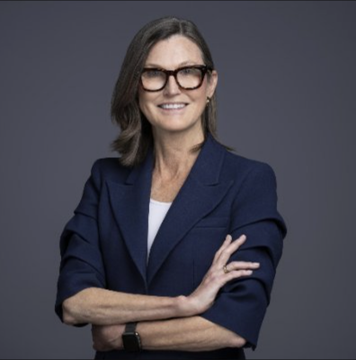 Cathie Wood, founder and CEO of Ark Invest, stands smiling with her arms crossed against a dark blue background, pictured in connection with her firm's recent purchase of 21,497 shares of Bullish ($BLSH) valued at $571,175.