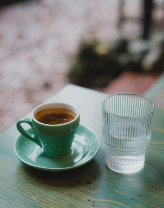 A small teal espresso cup filled with dark coffee sits beside a tall ribbed glass of water on a weathered green wooden table, with a blurred brick patio in the background. The cozy, relaxed café moment mirrors the post's vibe of winding down after payday and a long work week, enjoying a well-deserved break before the next grind.