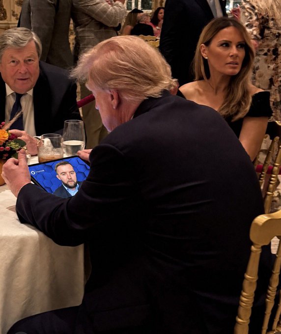 A man in a dark suit sits at a formal dinner table holding a tablet showing what appears to be a video call, while other well-dressed guests are seated nearby.