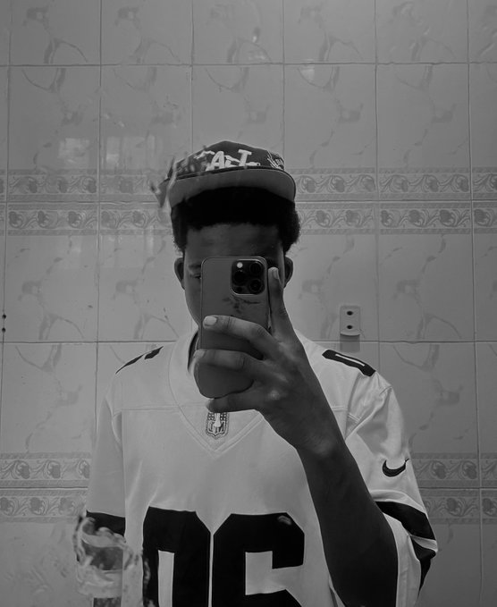 A black-and-white mirror selfie of a person wearing a cap and a sports jersey, holding a phone that obscures their face.